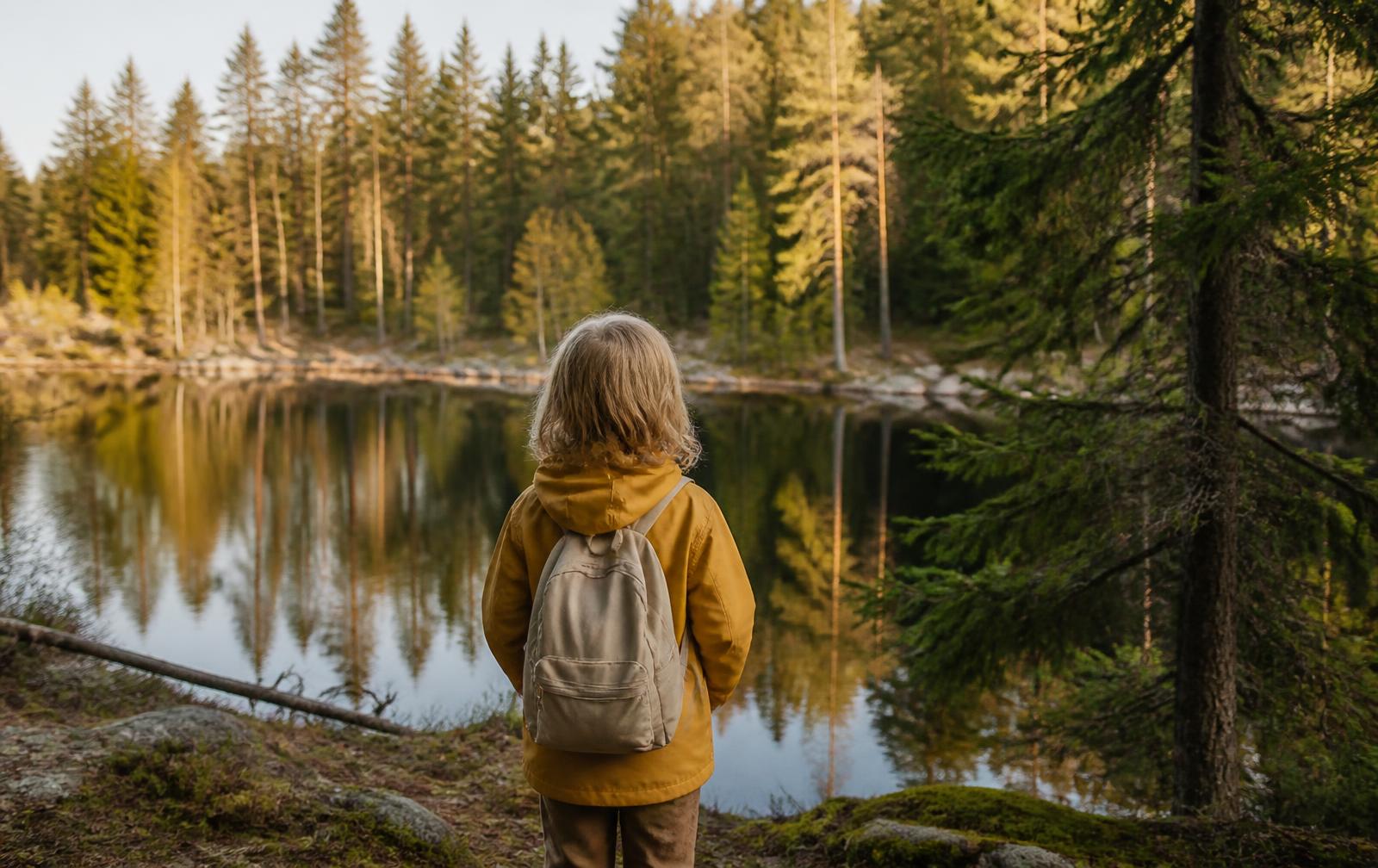 A child with a small backpack looking out over a forest lake